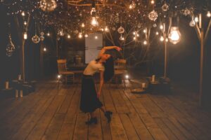 Uma mulher dança graciosamente sob lâmpadas quentes e iluminadas em um interior rústico de madeira.